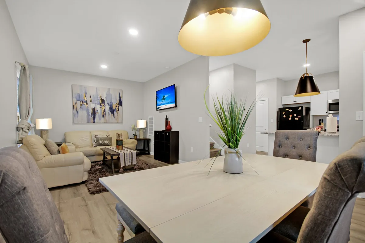 This interior shot presents a spacious open-plan living area with a dining table, a comfortable sofa, modern wall art, a television, and touches of greenery from indoor plants. The space is filled with natural light and arranged for both relaxation and social gatherings.