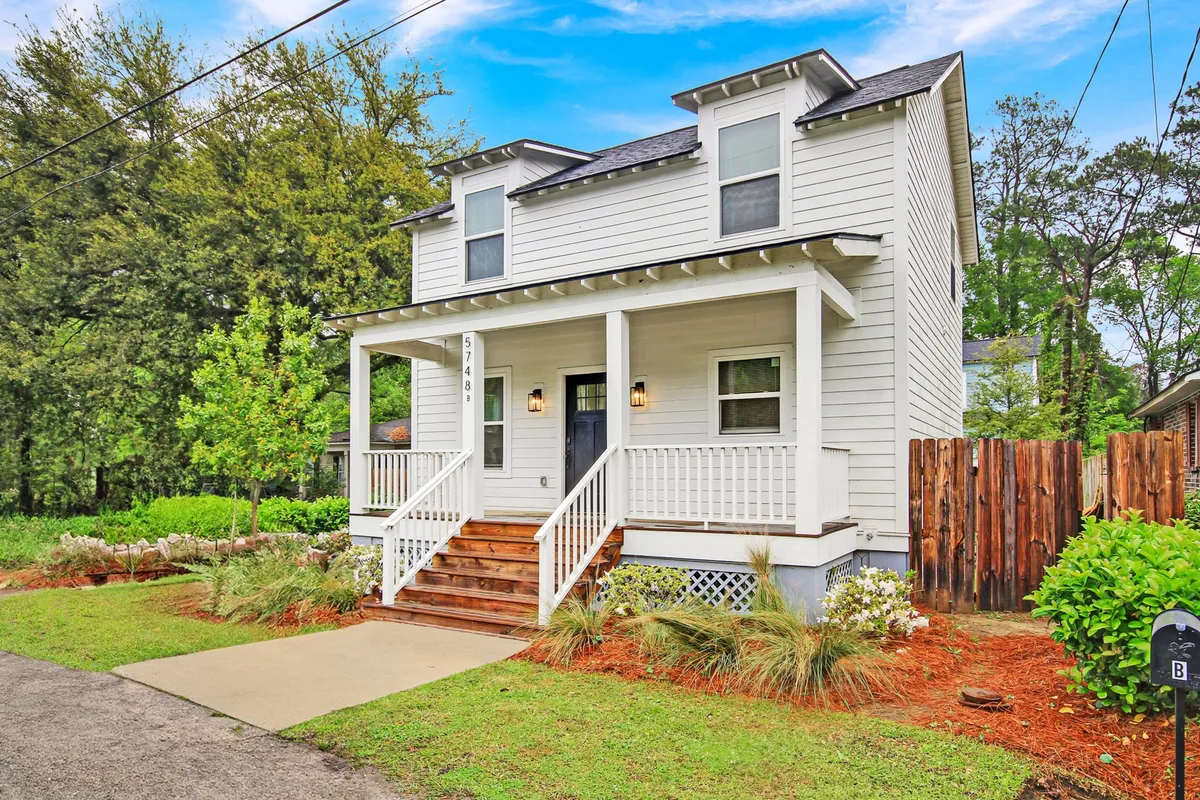 The image showcases the charming exterior of a new single-family home in Hanahan, South Carolina. A welcoming front porch with wooden stairs overlooks a beautifully landscaped and fenced yard, inviting guests to relax outdoors. The scene evokes the enjoyment of quiet mornings with coffee or lively evenings with friends and family, all surrounded by lush greenery.