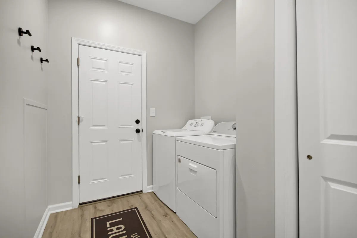A bright, tidy laundry room featuring a modern washer and dryer, a welcoming entry door, and a cozy laundry rug. The space is uncluttered and practical, clearly set up for easy laundry routines and organization of cleaning supplies.