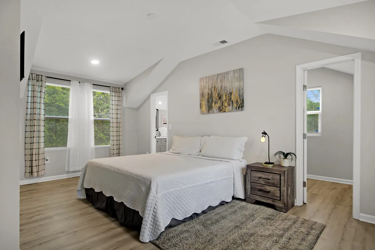 A tranquil bedroom with a plush bed, decorative pillows, a nightstand, contemporary artwork, and windows that allow gentle natural light to filter in. The uncluttered space conveys a sense of calm and personal sanctuary.