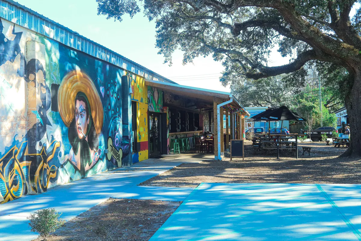 An outdoor dining area beneath a sprawling oak tree, surrounded by colorful graffiti murals. People gather at patio tables, sipping drinks and enjoying live music, while the creative atmosphere is heightened by vibrant street art and the presence of an outdoor bar.