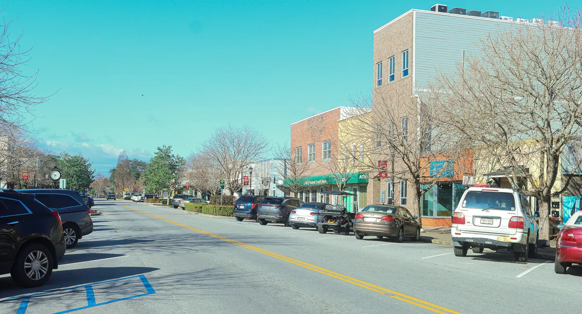 A lively streetscape featuring parked cars lining the road, storefronts with a mix of unique local businesses, and sidewalks shaded by trees. Small groups of people stroll, browse shops, and enjoy the vibrant street life, evoking the energy and communal feel of Park Circle.