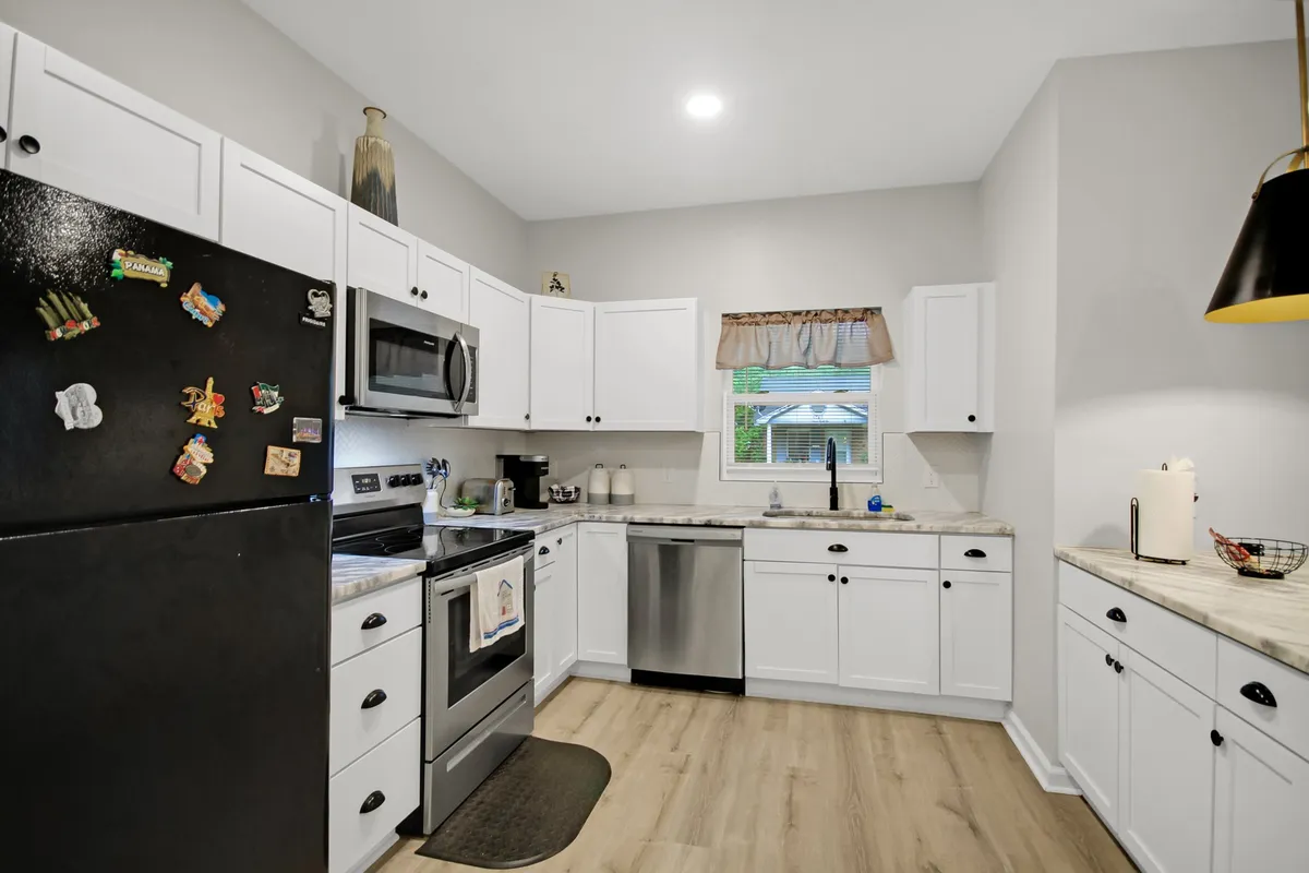 A modern kitchen with sleek white cabinetry, stainless steel appliances, a black refrigerator adorned with magnets, a marble countertop, and a kitchen sink overlooking a window. The space is cozy yet highly functional, ideal for meal prep or entertaining.