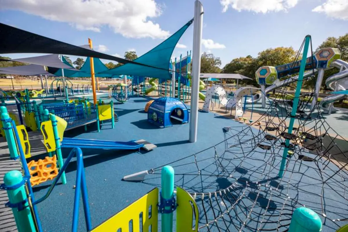 A vibrant outdoor playground with colorful play structures, slides, climbing nets, tunnels, and shade sails. Children are seen playing while parents supervise and groups gather around for picnics, all under a joyful atmosphere.
