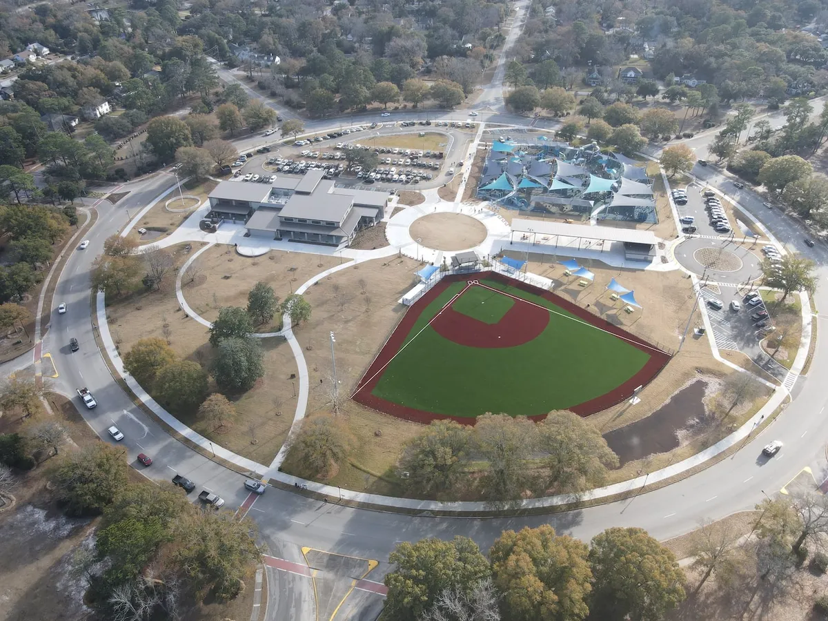 A community park scene featuring a baseball field, community center, and a prominent playground structure. Families are gathered around, children play, and picnic tables are set up under trees, highlighting the active community atmosphere.